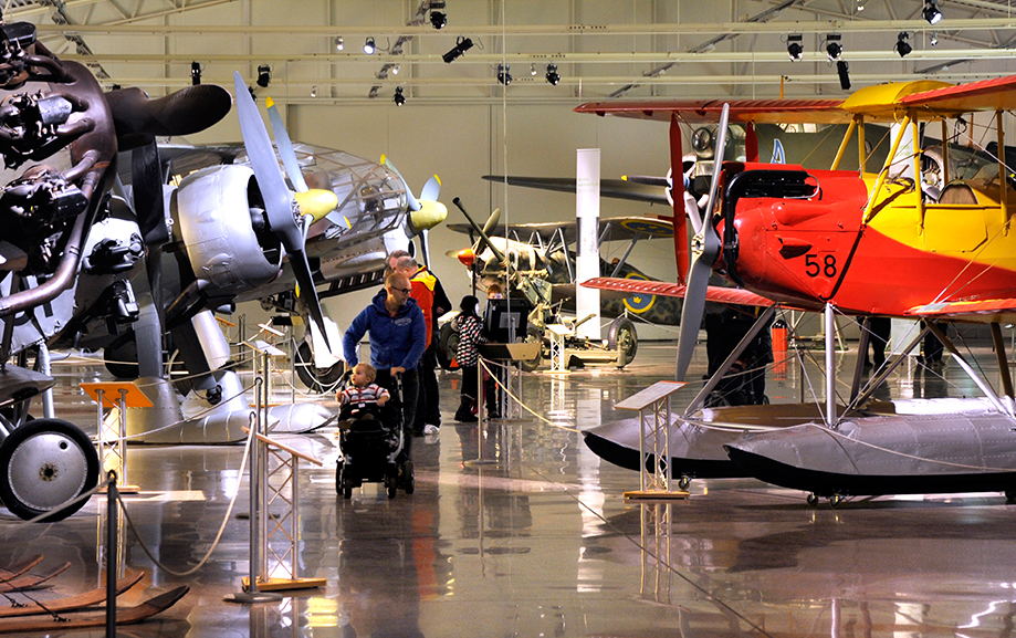 Bes&ouml;kare g&aring;r bland flygplanen i en av utst&auml;llningshallarna. Bland annat ett r&ouml;dvitt &auml;ldre plan med pontoner monterade f&ouml;r att kunna g&aring; p&aring; vatten.