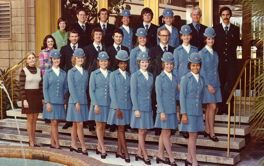 Flygv&auml;rdinnor och kabinpersonal i ljusbl&aring; uniformer och hattar. Personerna &auml;r uppst&auml;llda i flera rader framf&ouml;r en byggnad med glasd&ouml;rrar och guldramade f&ouml;nster.