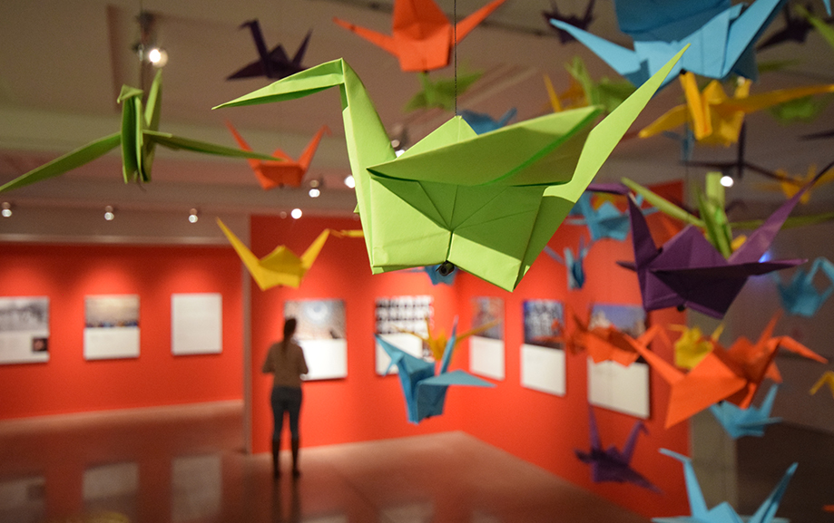 En n&auml;rbild p&aring; en gr&ouml;n origamitrana i papper i taket, i bakgrunden ser man fler tranor samt en bes&ouml;kare som kollar in utst&auml;llningen av tavlor.