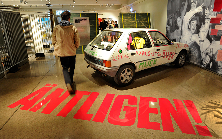 En bes&ouml;kare g&aring;r igenom ett rum d&auml;r det st&aring;r "&Auml;ntligen" i stora r&ouml;da bokst&auml;ver p&aring; golvet. Bredvid st&aring;r en bil parkerad med text m&aring;lad p&aring; sig: "Muck", "&Auml;ntligen &auml;r skiten &ouml;ver" samt ett nummer.