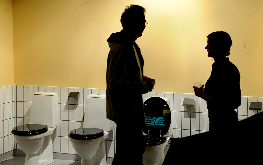 Tv&aring; personer st&aring;r som svarta siluetter i ett halvkaklat badrum med toaletter p&aring; rad bredvid varandra. Varje toalett har en svart sits och en tillh&ouml;rande toalettpappersh&aring;llare bredvid sig.