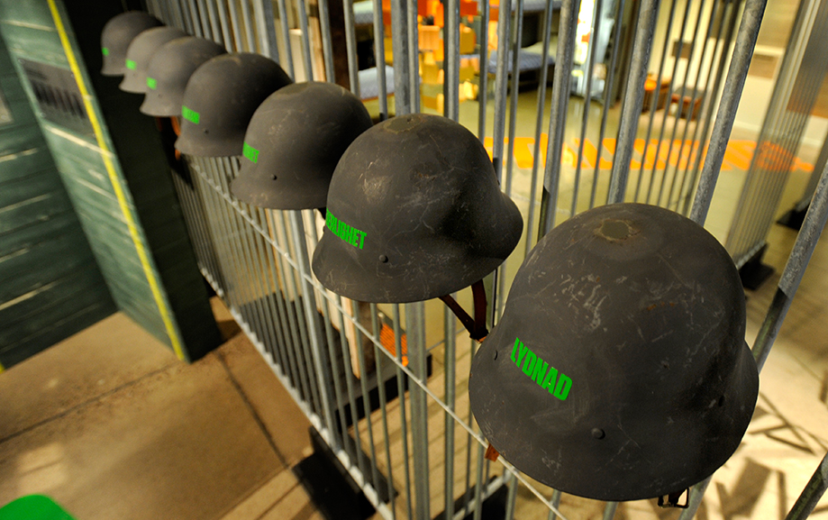 Svarta skyddshj&auml;lmar p&aring; rad mot ett galler. Bakp&aring; hj&auml;lmarna st&aring;r ett ord i gr&ouml;n text. Den n&auml;rmsta hj&auml;lmen har texten "Lydnad" p&aring; sig.