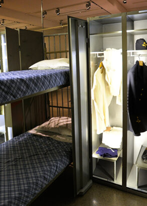 En sovsal med v&auml;lb&auml;ddade v&aring;ningss&auml;ngar med bl&aring;rutiga lakan och vita kuddar, Mellan tv&aring; s&auml;ngar ett pl&aring;tsk&aring;p med skor och kl&auml;der som anv&auml;ndes i lumpen.