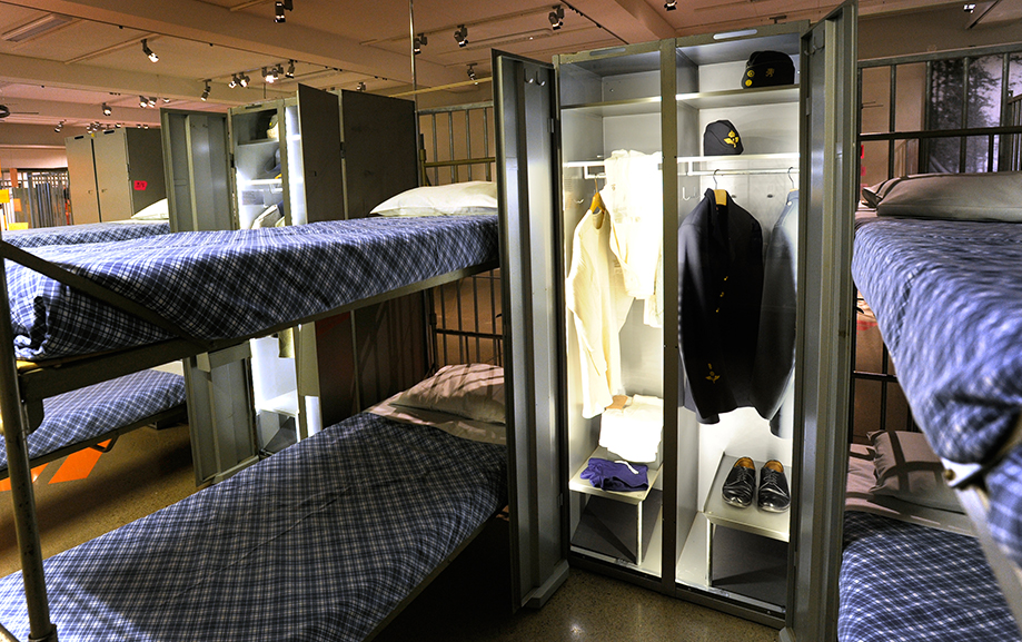 En sovsal med v&auml;lb&auml;ddade v&aring;ningss&auml;ngar med bl&aring;rutiga lakan och vita kuddar, Mellan tv&aring; s&auml;ngar ett pl&aring;tsk&aring;p med skor och kl&auml;der som anv&auml;ndes i lumpen.