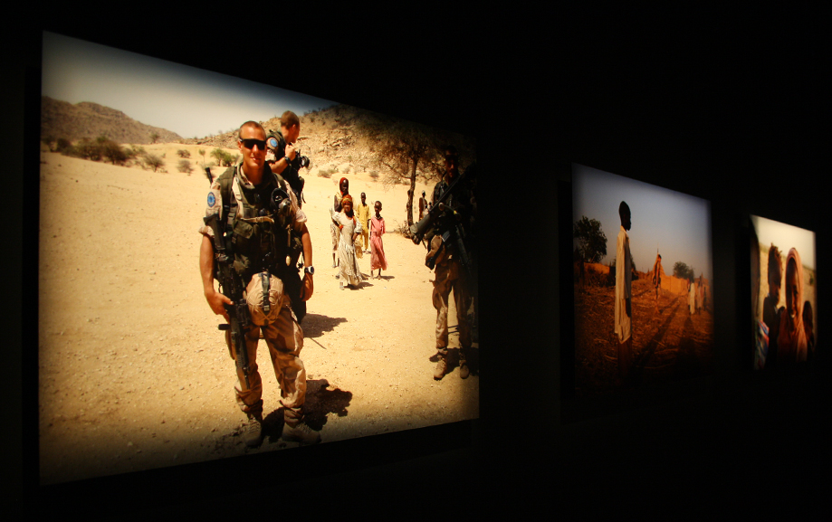 Foton som h&auml;nger mot en svart v&auml;gg, det i f&ouml;rgrunden f&ouml;rest&auml;ller soldater som vandrar genom &ouml;knen och tr&auml;ffar p&aring; civila.