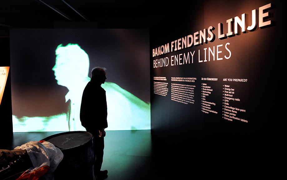 En bes&ouml;kare st&aring;r som en siluett i ett svagt upplyst rum framf&ouml;r informationstavlan "Bakom fiendens linje".