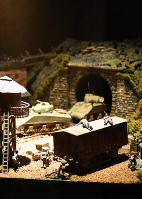 Ett modelbygge &ouml;ver ett utkikstorn med balkong runtom med stege upp, nedanf&ouml;r en stuga och i bakgrunden tv&aring; gr&ouml;na stridsvagnar som kommer ut ur en t&aring;gtunnel i berget.