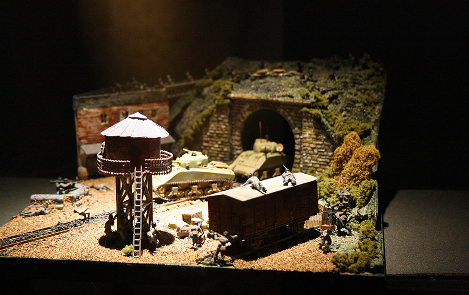 Ett modelbygge &ouml;ver ett utkikstorn med balkong runtom med stege upp, nedanf&ouml;r en stuga och i bakgrunden tv&aring; gr&ouml;na stridsvagnar som kommer ut ur en t&aring;gtunnel i berget.