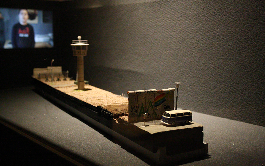 En bl&aring;vit folkabuss bakom en h&ouml;g mur i f&ouml;rgrunden och lite l&auml;ngre bort ett flygtorn p&aring; ett flygf&auml;lt. Bakom byggmodellen skymtar en p&aring;g&aring;ende filmvisning.