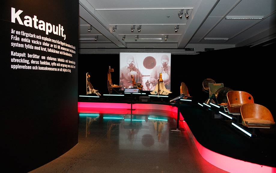 P&aring; ena v&auml;ggen st&aring;r det Katapult i vitt mot svart bakgrund och sedan f&ouml;ljer text om katapultstolens historia. Strax intill st&aring;r flygplansstolar p&aring; rad och visar p&aring; utvecklingen fr&aring;n enklaste tr&auml;stol till de mer avancerade stolar vi k&auml;nner igen i dag.