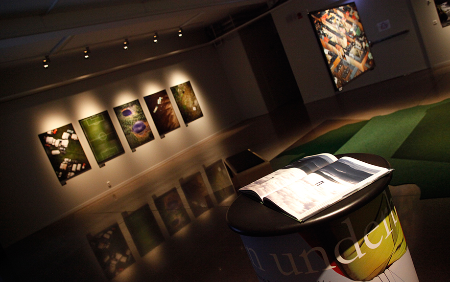 En dovt upplyst utst&auml;llningssal med diverse bilder av Sverige p&aring; v&auml;ggarna, d&auml;ribland en gr&ouml;n fotbollsplan sedd fr&aring;n ovan.