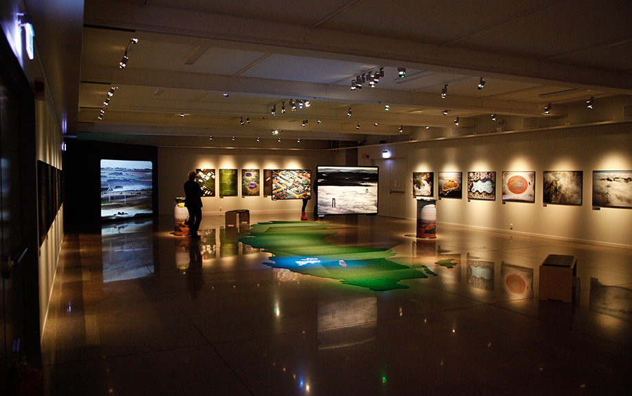 En gr&ouml;nbl&aring; matta i form av Sverige p&aring; golvet i en utst&auml;llningshall med tavlor och informationsplaketter p&aring; v&auml;ggarna. En bes&ouml;kare som besk&aring;dar utst&auml;llningen.