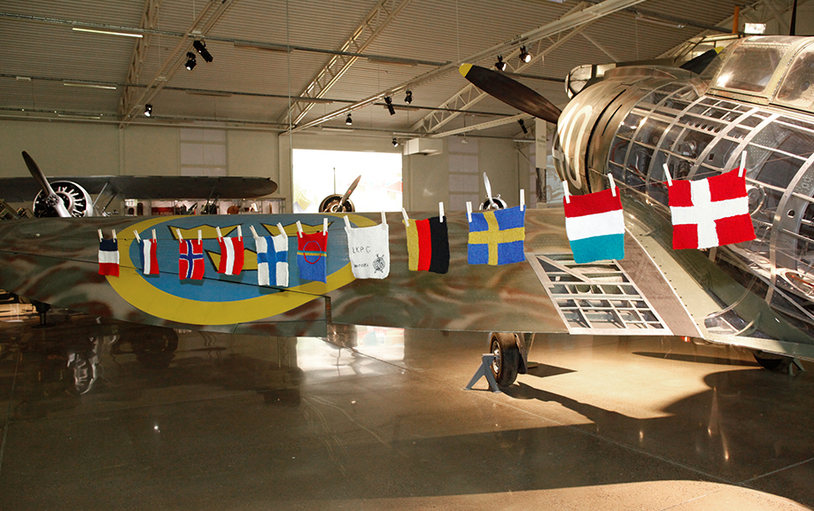 Stickade landsflaggor upph&auml;ngda med kl&auml;dnypor p&aring; lina h&auml;nger mellan tv&aring; flygplan i en utst&auml;llningshall.