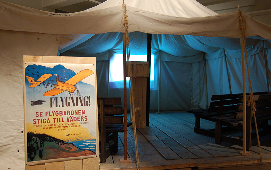 Ett vitt t&auml;lt med tr&auml;b&auml;nkar uppst&auml;llda p&aring; rad inuti. Utanf&ouml;r ing&aring;ngen st&aring;r en skylt med texten "Flygning. Se flygbaronen stiga till v&auml;ders"