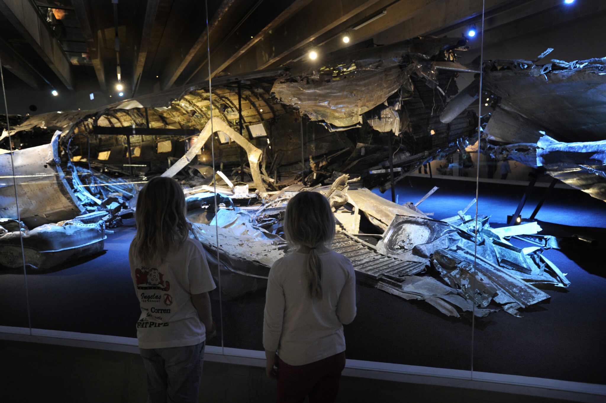 Tv&aring; barn st&aring;r i en sal med d&auml;mpat ljus och tittar p&aring; den b&auml;rgade DC3:an som ligger i en stor glasmonter.