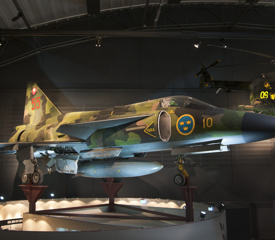 Stridsflygplanet Saab 37 Viggen i svensk kamouflagef&auml;rg, utst&auml;lld p&aring; museum. Planet &auml;r m&auml;rkt med siffran 10 och det svenska flygvapnets symbol med tre kronor. P&aring; fenan syns nummer 55 i r&ouml;tt. I bakgrunden skymtar en milit&auml;rhelikopter med siffran 09