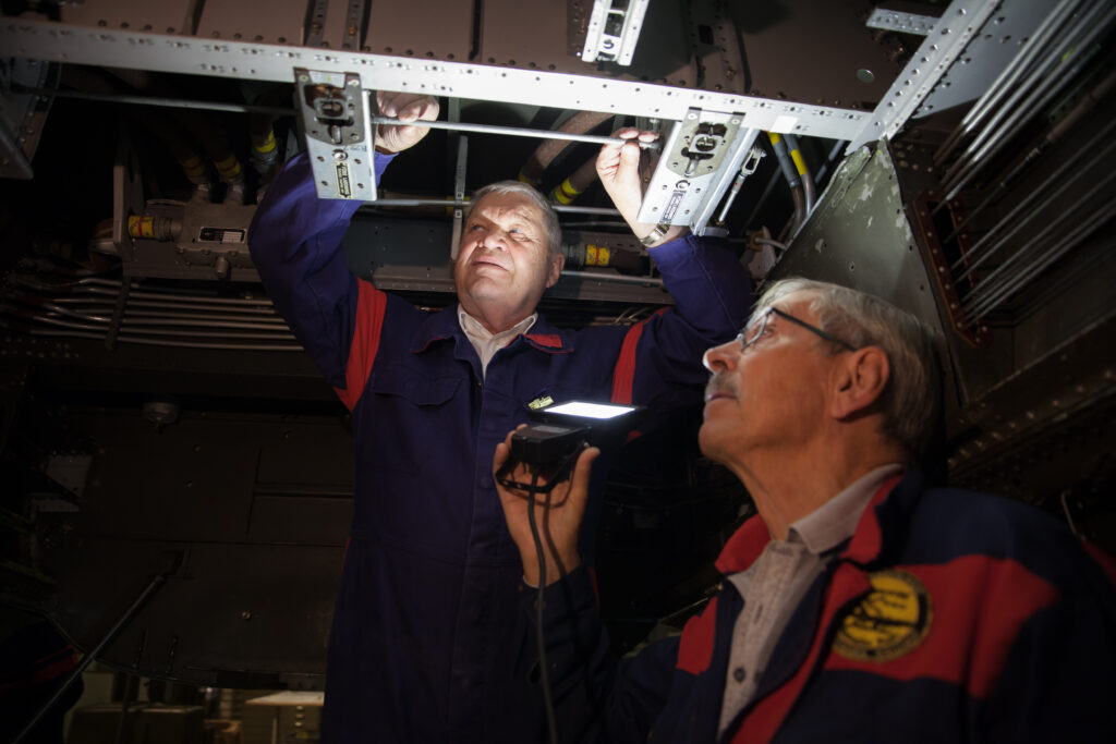 Två volontärer inspekterar under ett flygplan. den ena håller upp en ficklampa så den andra kan se bättre.