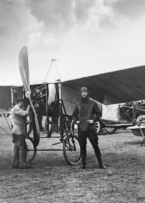 Carl Cederstr&ouml;m st&aring;r, flygredo med pilotm&ouml;ssa p&aring;, framf&ouml;r ett flygplan p&aring; ett f&auml;lt. Bredvid st&aring;r en man som kollar motorn. I bakgrunden syns &aring;sk&aring;dare.