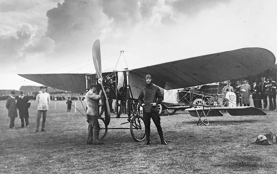 Carl Cederstr&ouml;m st&aring;r, flygredo med pilotm&ouml;ssa p&aring;, framf&ouml;r ett flygplan p&aring; ett f&auml;lt. Bredvid st&aring;r en man som kollar motorn. I bakgrunden syns &aring;sk&aring;dare.