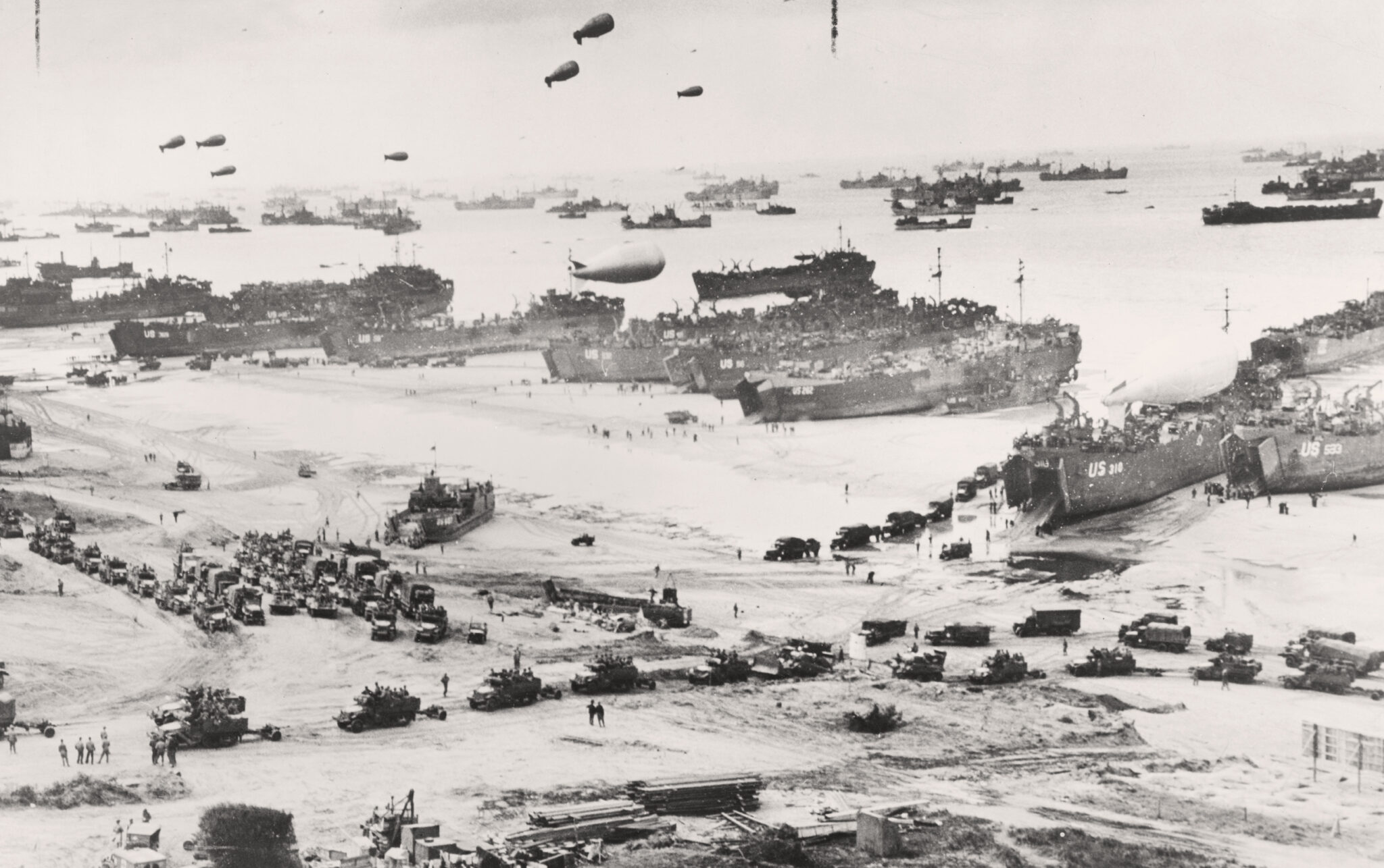 Svartvit historisk bild av Normandiestranden under D-dagen 1944. Stora landstigningsfartyg ligger vid stranden medan m&auml;ngder av milit&auml;rfordon, soldater och utrustning r&ouml;r sig i land. Himlen fylls av f&ouml;rankrade ballonger och horisonten av fartyg.