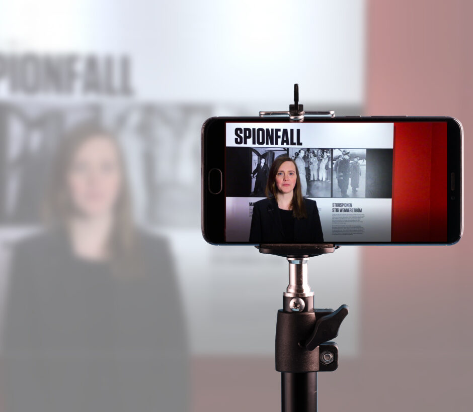 En mobilkamera h&aring;lls upp i ett stativ f&ouml;r att spela in inneh&aring;ll till digital visning, en kvinna st&aring;r framf&ouml;r kameran och bakom henne text till utst&auml;llningen Spionfall