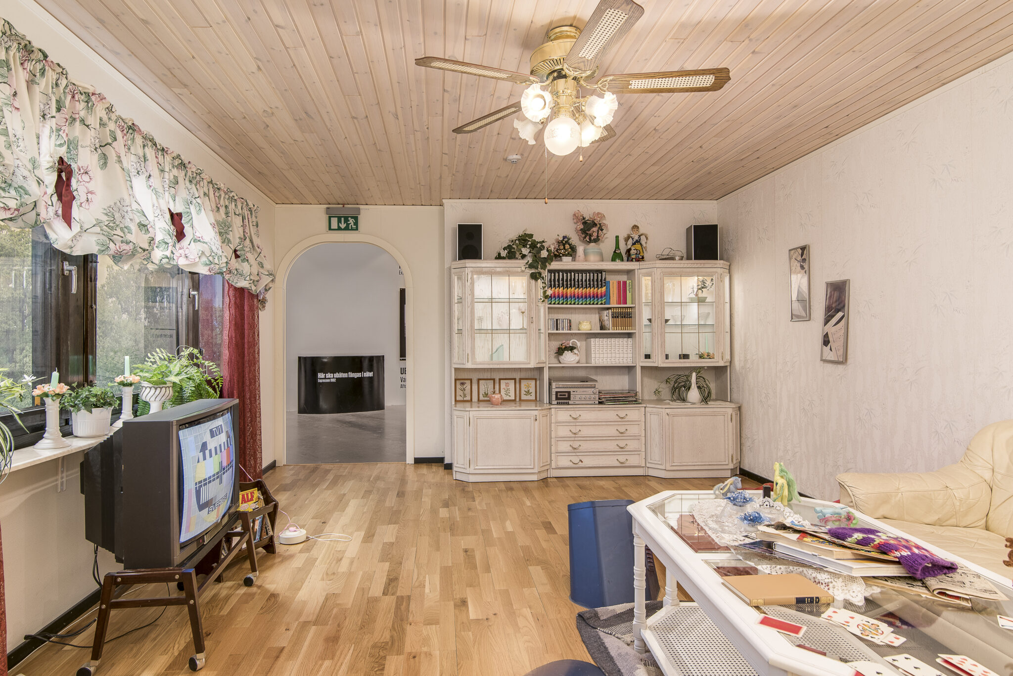 Ett vardagsrum som h&auml;mtat ur det svenska folkhemmet med takfl&auml;kt, tv, skinnsoffa, vitt soffbord och blommiga gardiner. I bakgrunden en typisk vardagsrumsm&ouml;bel f&ouml;r prydnadssaker, fotografier och b&ouml;cker med mera.