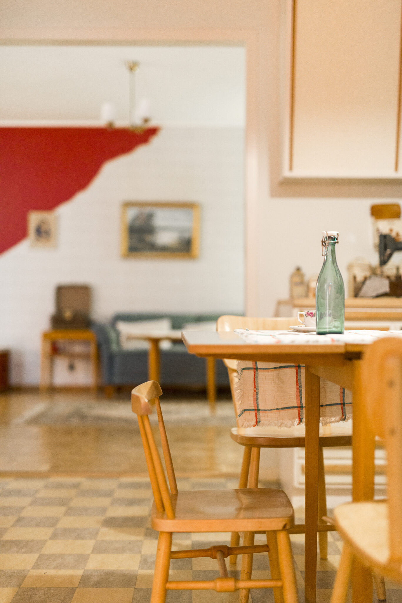 Folkhemsmilj&ouml;, ett tr&auml;bord med duk och pinnstolar omkring i f&ouml;rgrunden och ett vardagsrum med soffa, soffbord, tavla p&aring; v&auml;ggen och en taklampa med glaskupor i bakgrunden.