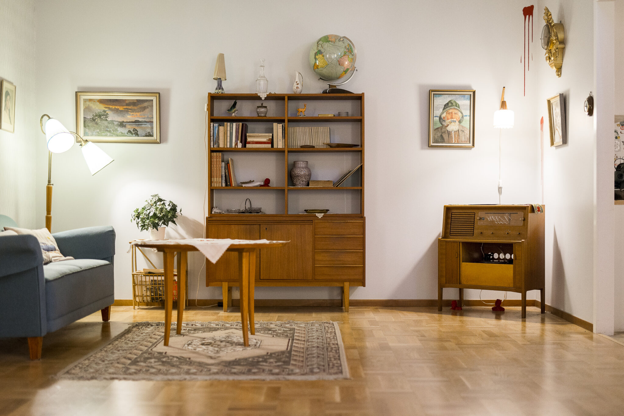 Ett vardagsrum inrett med teak - bokylla, radiom&ouml;bel, soffbord, golvlampa och soffa med bl&aring;tt tyg. uppep&aring; bokhyllan st&aring; en jordglob och p&aring; v&auml;ggarna h&auml;nger tavlor och ett svulstigt v&auml;ggur med bladguld.