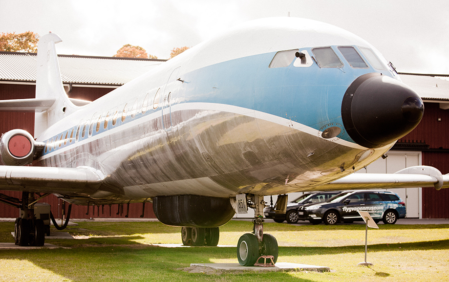 Ett bl&aring;vitt jetflyglan st&aring;endes p&aring; marken framf&ouml;r ett r&ouml;tt skjul, d&auml;r det st&aring;r n&aring;gra bilar parkerade. Planet heter Sud Aviation Caravelle och har motorerna placerade i akterpartiet.