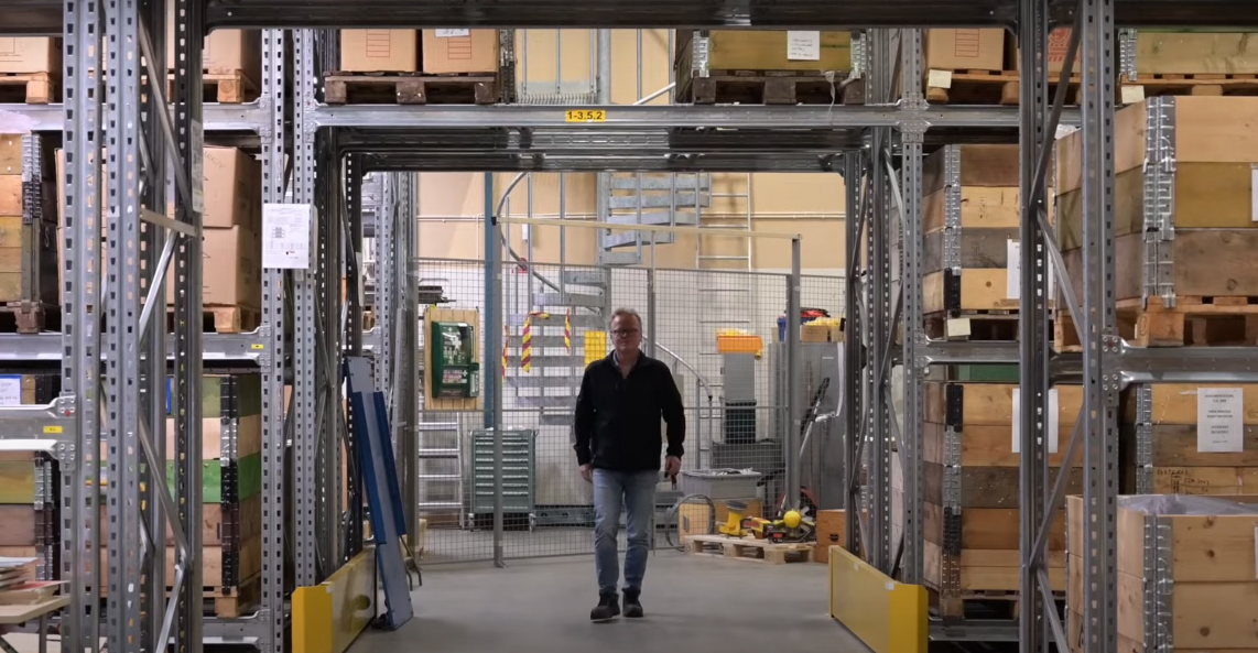 En person st&aring;r i mitten av ett f&ouml;rr&aring;d, hyllor l&auml;ngs sidorna med l&aring;dor p&aring;, en spiraltrappa i st&aring;l i bakgrunden.