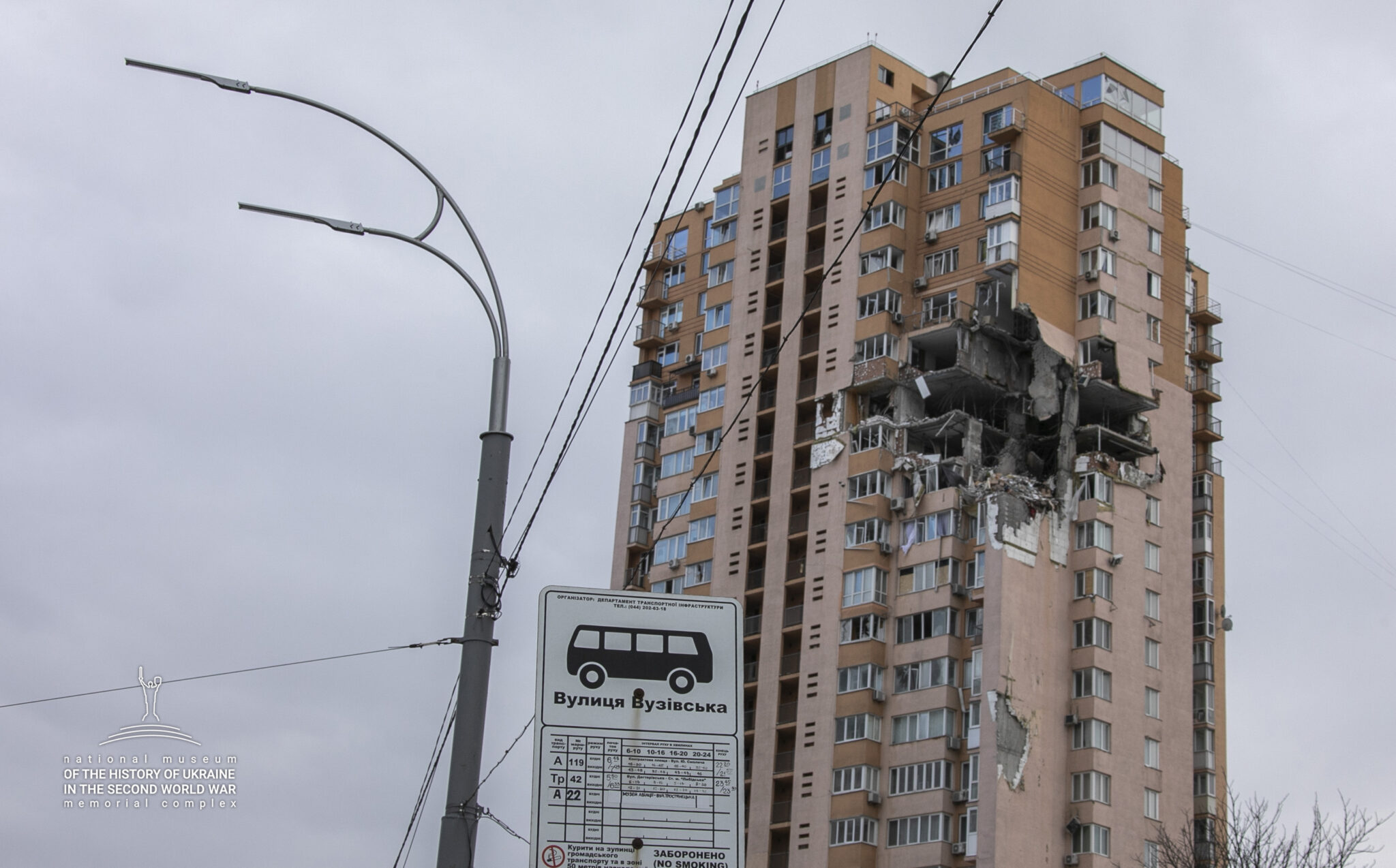 Ett h&ouml;ghus som har ett stort h&aring;l i ena sidan d&auml;r det troligtvis har farit in en bomb. I f&ouml;rgrunden en bussh&aring;llplatsskylt.