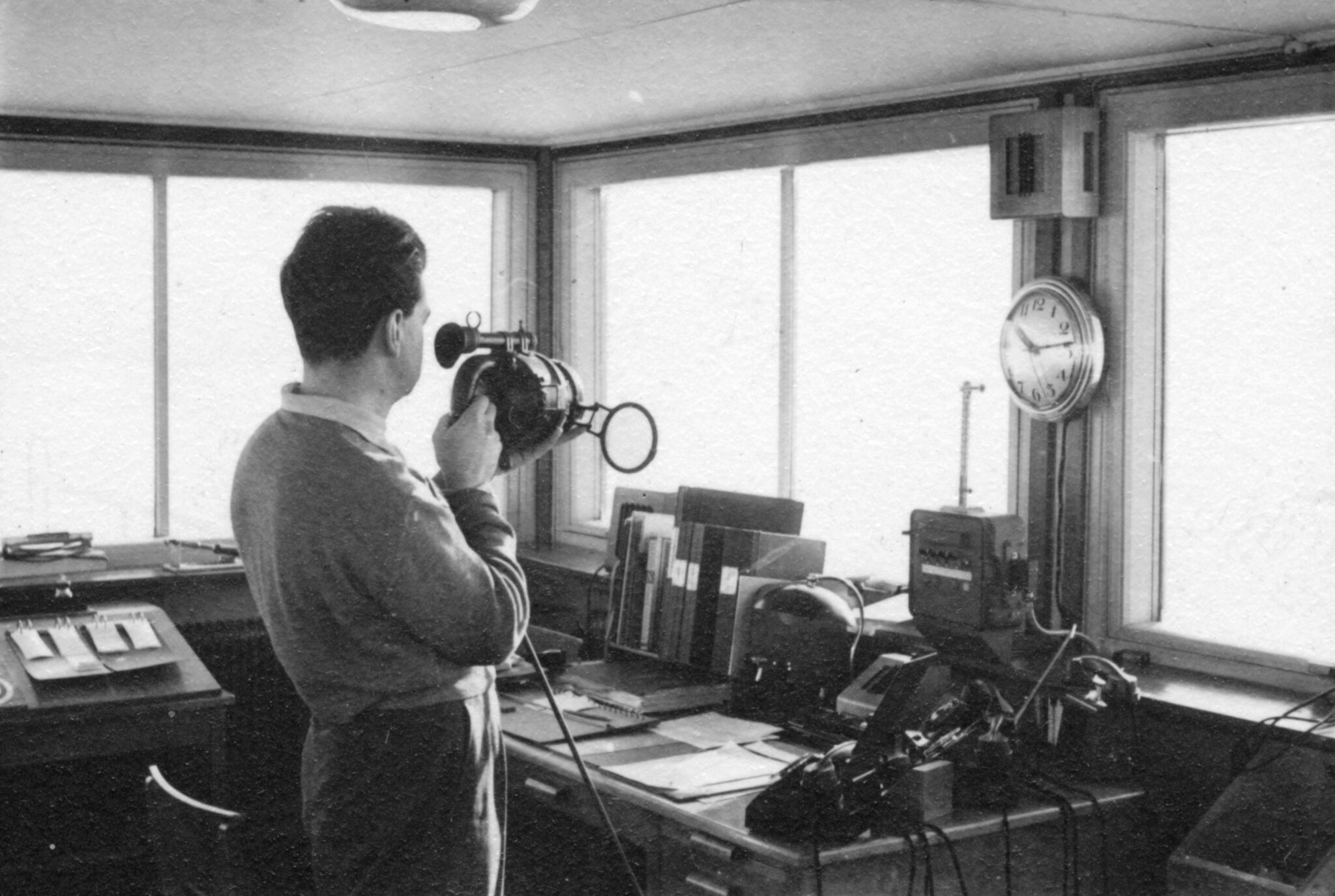 En svartvit bild ur arkivet p&aring; en person som st&aring;r i ett trafikledartorn, blickandes ut genom f&ouml;nstret och signalerar med signallampa.