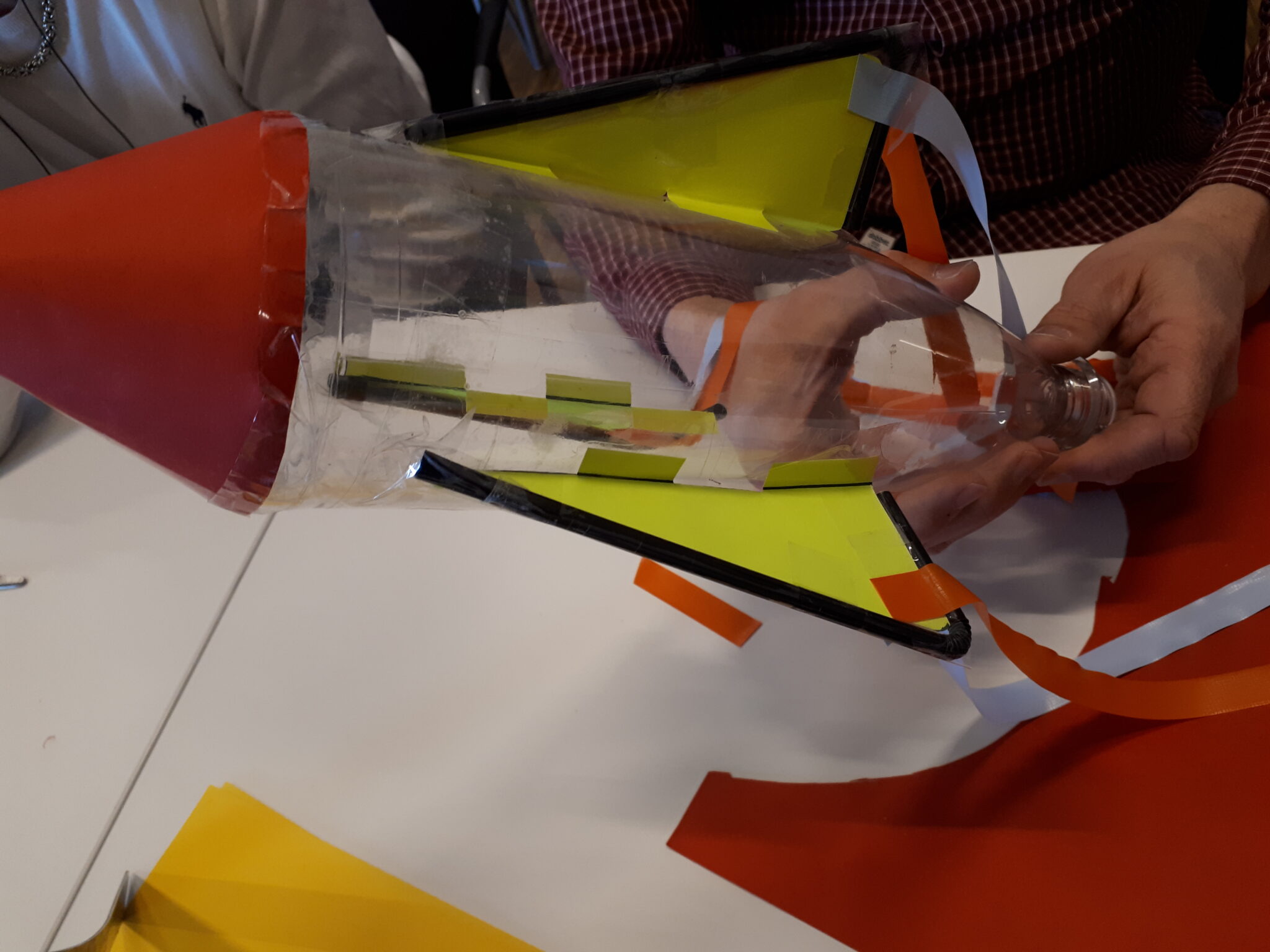 En person h&aring;ller i en hemmagjord rymdraket byggd av en petflaska, f&auml;rgat papper och tejp. Raketen har fenor i neongult och svart samt en r&ouml;d konformad spets. Pysselmaterial ligger utspritt p&aring; bordet.