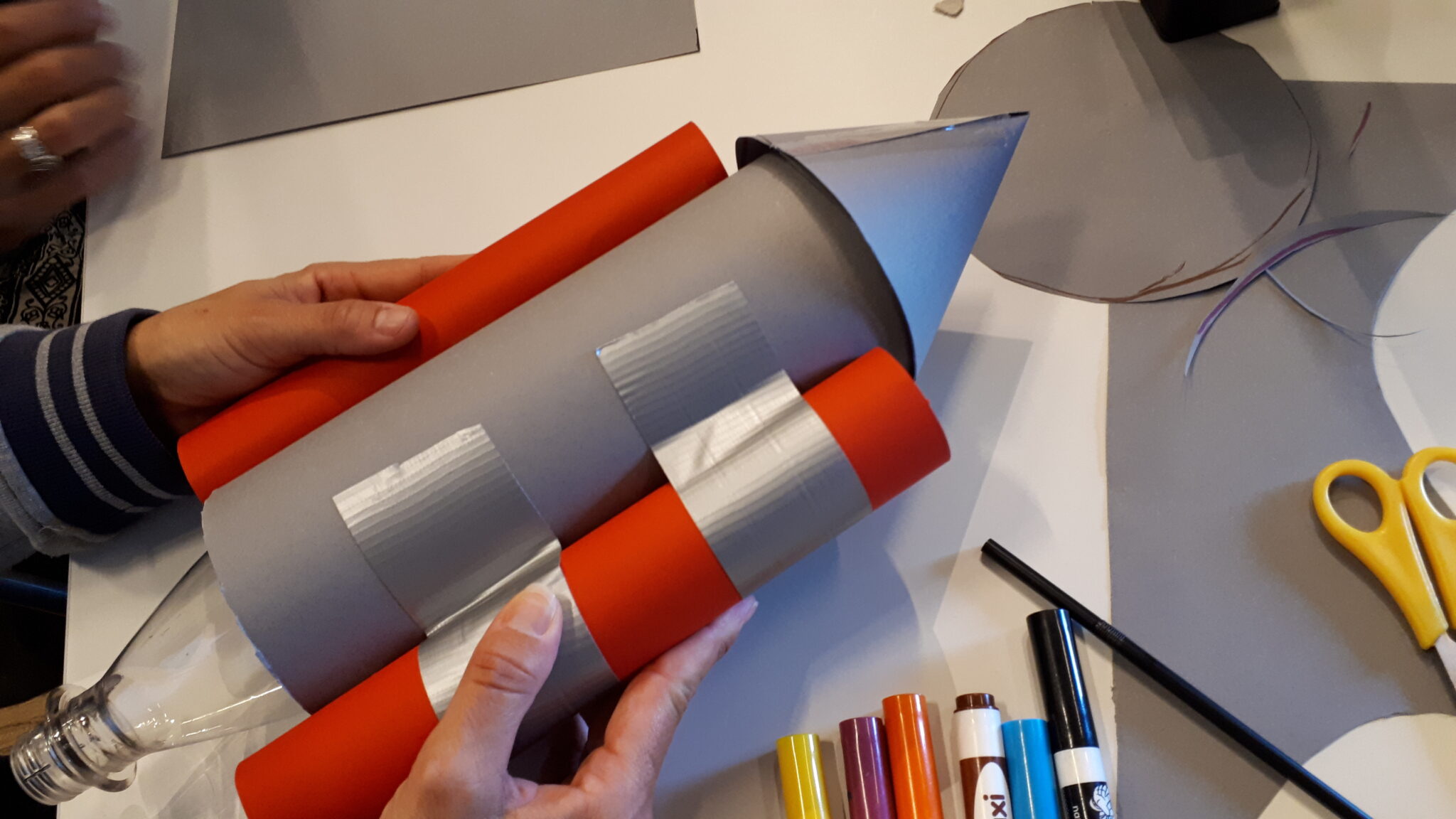 Tv&aring; barnh&auml;nder h&aring;ller i en raket byggd av papper, ihoptejpad med silvertejp.