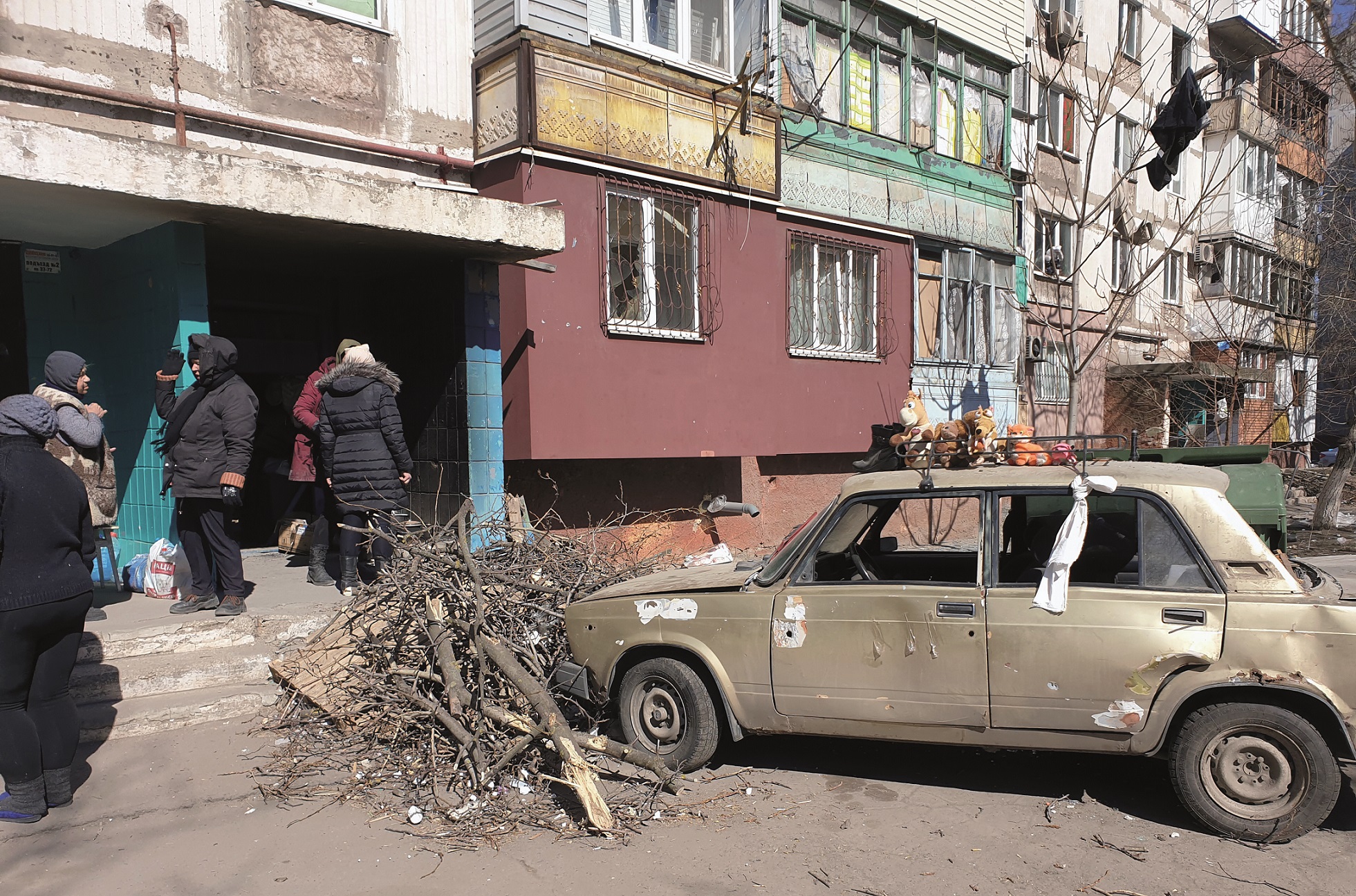 En silverf&auml;rgad nedg&aring;ngen bil st&aring;r med f&ouml;ren ink&ouml;rd i en rish&ouml;g framf&ouml;r ett flerfamiljshus. Framf&ouml;r entr&eacute;n st&aring;r fem personer i olika grupperingar.