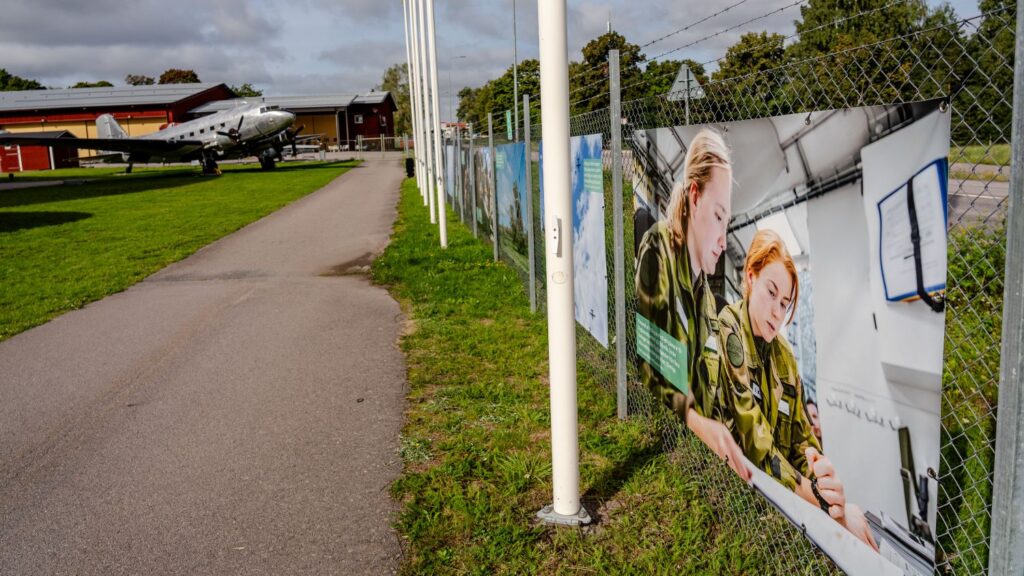 En utställning utomhus längs ett gunnebostaket och ett silverfärgat flygplan bredvid. Närmsta bilden visar två personer i grön kamouflage, 