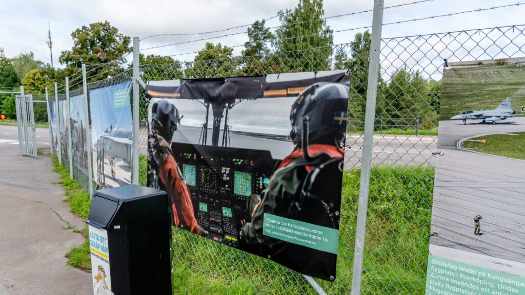 En utställning utomhus med flera fotografier uppsatta längs ett gunnebostaket. Den i fokus här föreställer två personer i en cockpit som flyger över vatten. 
