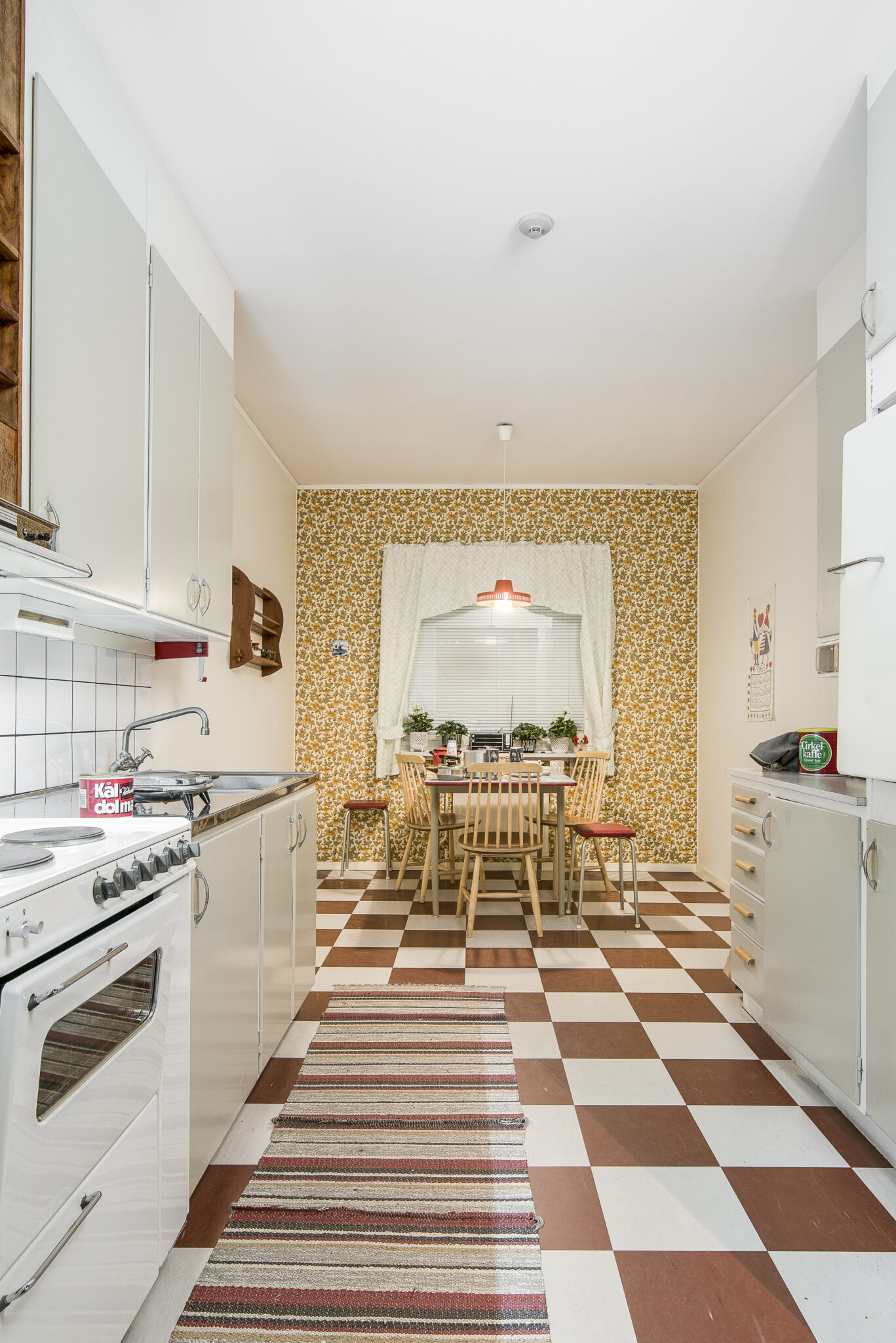 Ett klassiskt funkisk&ouml;k med brunvitrutigt golv, tr&auml;bord med pinnstolar runtom en orange taklampa och en gulblommig fondv&auml;gg.