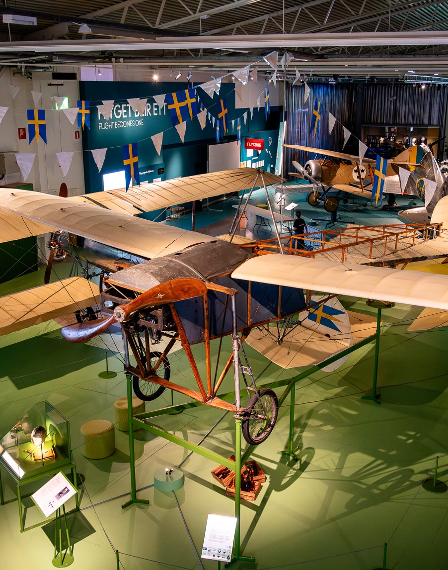 Ett &auml;ldre propellerflygplan till stor del tillverkat i tr&auml; med vingar kl&auml;dda med tyg. St&aring;r i en utst&auml;llningshall med fler plan skymtandes bakom.