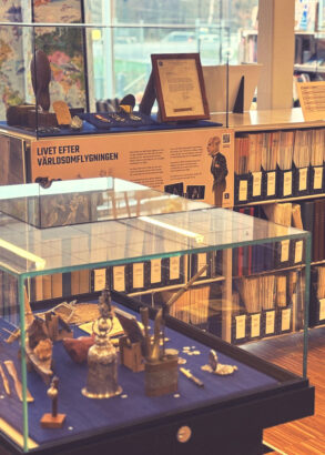 En glasmonter med diverse souvenirer mot en bl&aring; botten. I bakgrunden bokhylla med fulla dokumenth&aring;llare.