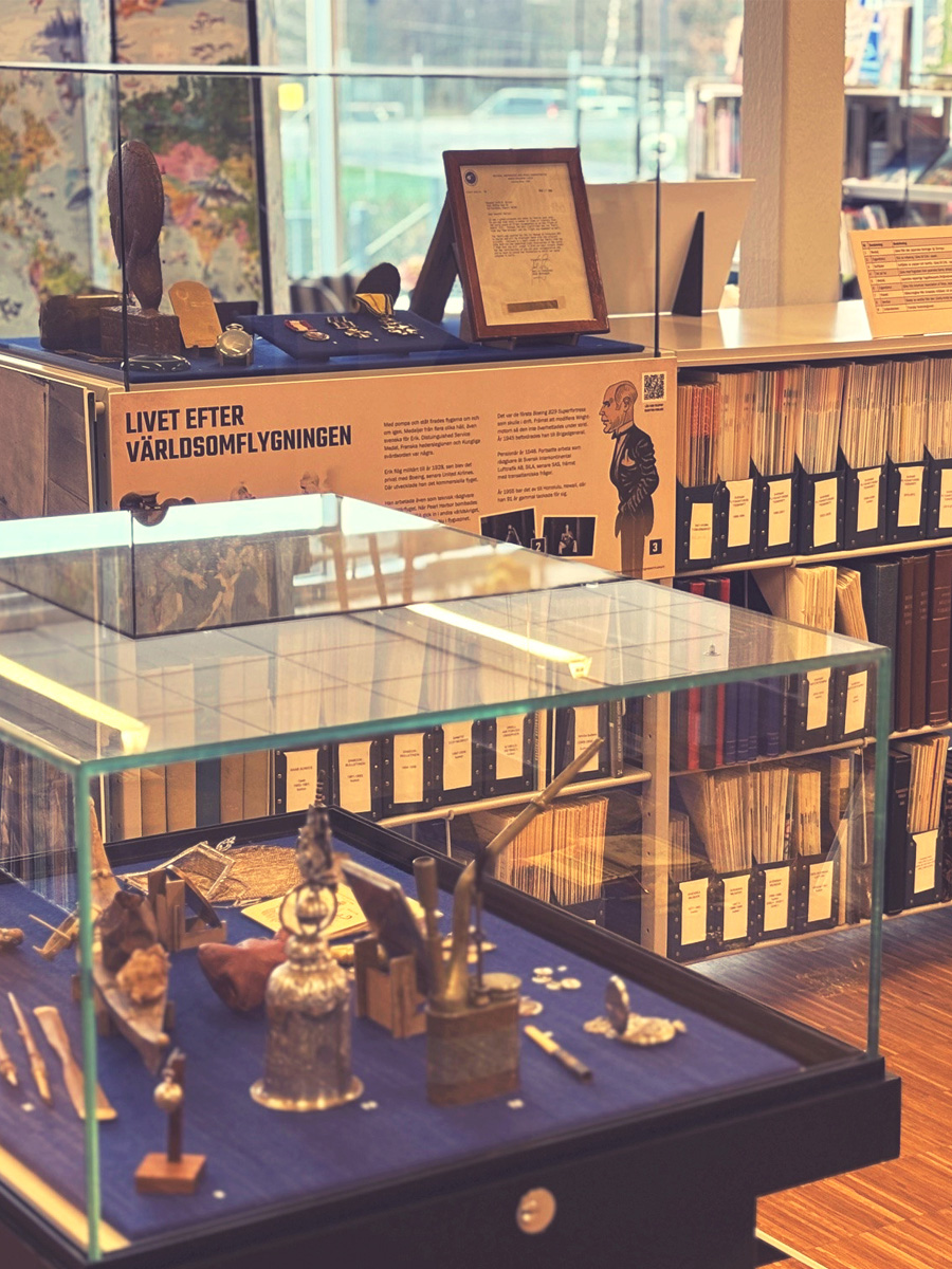 En glasmonter med diverse souvenirer mot en bl&aring; botten. I bakgrunden bokhylla med fulla dokumenth&aring;llare.