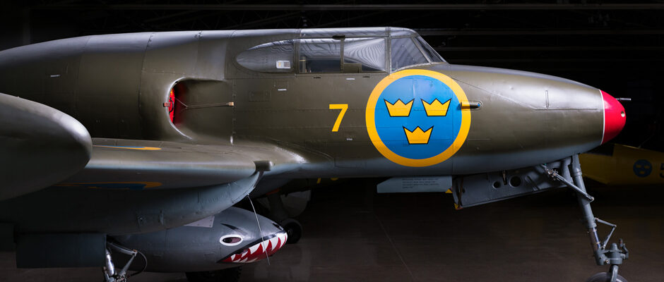 N&auml;rbild p&aring; en svensk milit&auml;rflygplansnos med det svenska flygvapnets bl&aring;gula rundel med tre kronor, siffran 7 i gult och en hajmun-m&aring;lning p&aring; vapenkapseln under vingen. Flygplanet st&aring;r inomhus.